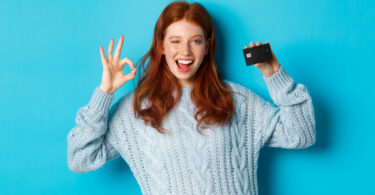 Happy redhead girl in sweater showing credit card and okay sign, recommending bank offer, standing over blue background