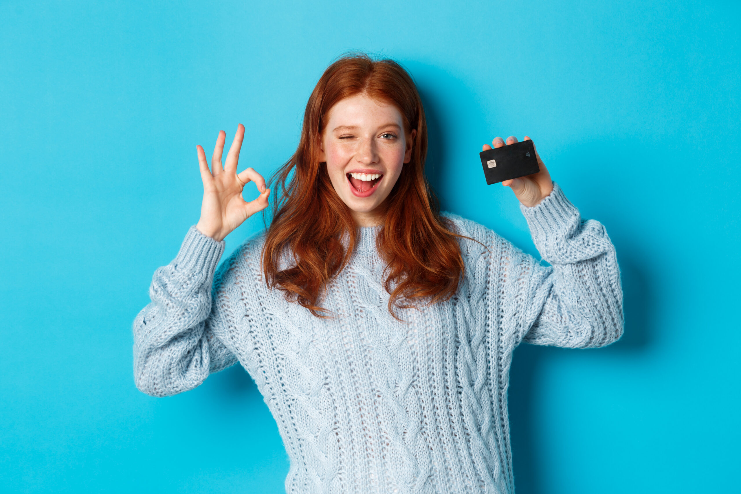 Happy redhead girl in sweater showing credit card and okay sign, recommending bank offer, standing over blue background